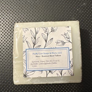 Puede incluir: Una pastilla de jabón cuadrada envuelta en plástico transparente. La etiqueta blanca tiene el texto "Fluffy Cow Soaps & More, LLC Non-Scented Beef Tallow" y un diseño floral. Los ingredientes están listados debajo.