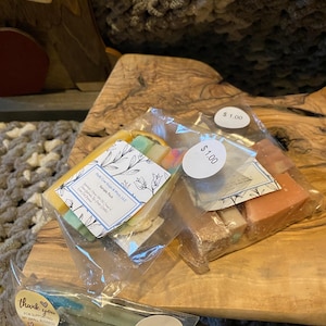 Puede incluir: Tres bolsas de barras de jabón de colores con una etiqueta de precio de $ 1.00 en cada bolsa. Las barras de jabón están envueltas en plástico transparente y tienen una variedad de colores y patrones. Las bolsas están sobre una superficie de madera. El texto en el empaque del jabón dice "PlyC Spears, LLC Sample Pack".