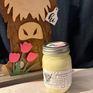 Op de afbeelding: Een pot Dandelion & Beef Tallow zonnebrandcrème met een metalen deksel staat voor een houten koeienuitsparing met roze tulpen. De pot heeft een etiket met tekst en een koe-illustratie. De zonnebrandcrème is ongeveer 340g.