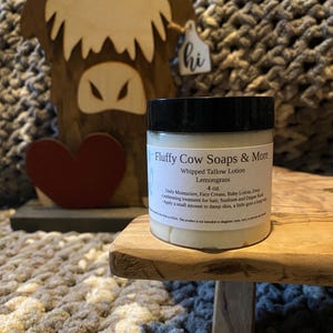 May include: A white jar with a black lid labeled "Fluffy Cow Soaps & More Whipped Tallow Lotion Lemongrass 4 oz. Daily Moisturizer, Face Cream, Baby Lotion, Deep conditioning treatment for hair. Sunburn and Diaper Rash - Apply a small amount to damp skin, a little goes a long way."