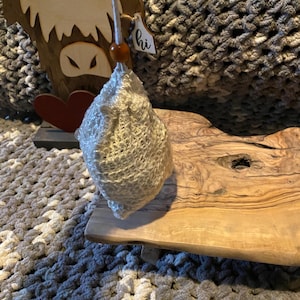 May include: A decorative wooden Highland cow with a red heart and a small tag that says "hi". A light-colored, textured item is suspended from the cow. The cow is on a wooden surface, with a gray and white textured blanket in the background.