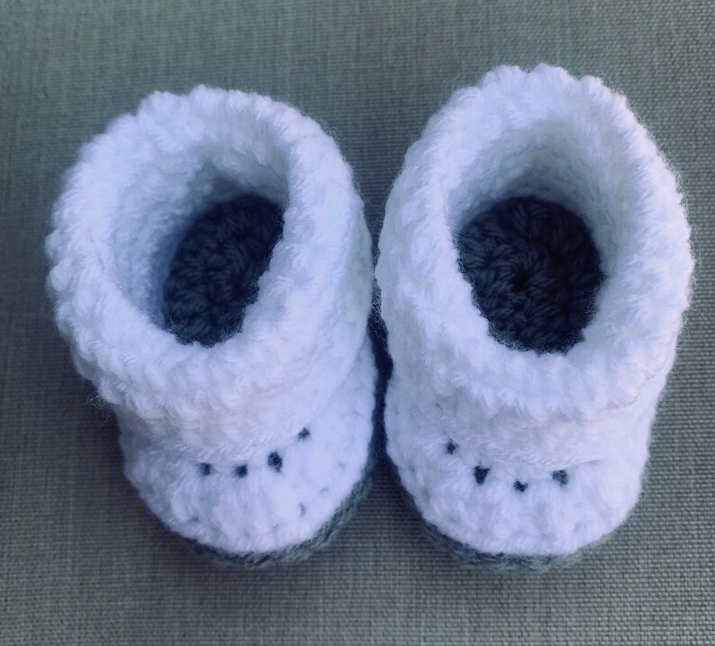 Op de afbeelding: Een paar witte gehaakte babyschoentjes met grijze gehaakte zolen.