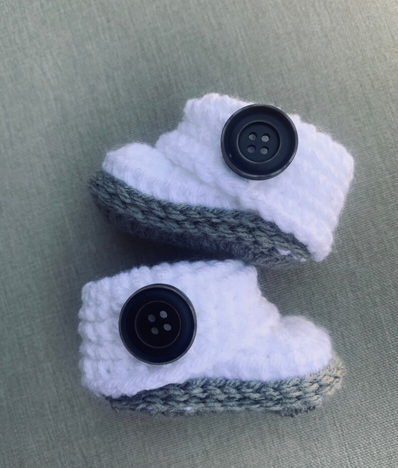 Op de afbeelding: Een paar witte gebreide babyschoentjes met grijze bies en zwarte knopen.