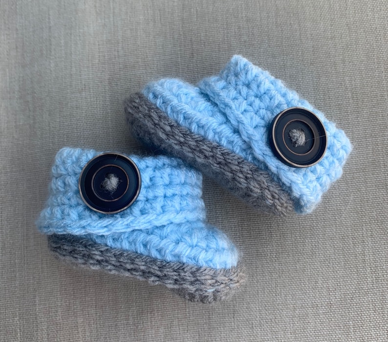 Op de afbeelding: Een paar babyschoentjes in lichtblauw met grijze randjes en zwarte knopen.