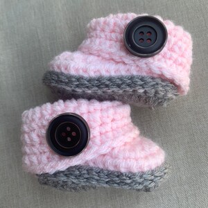 Op de afbeelding: Een paar roze en grijze gehaakte babyschoentjes met zwarte knopen.