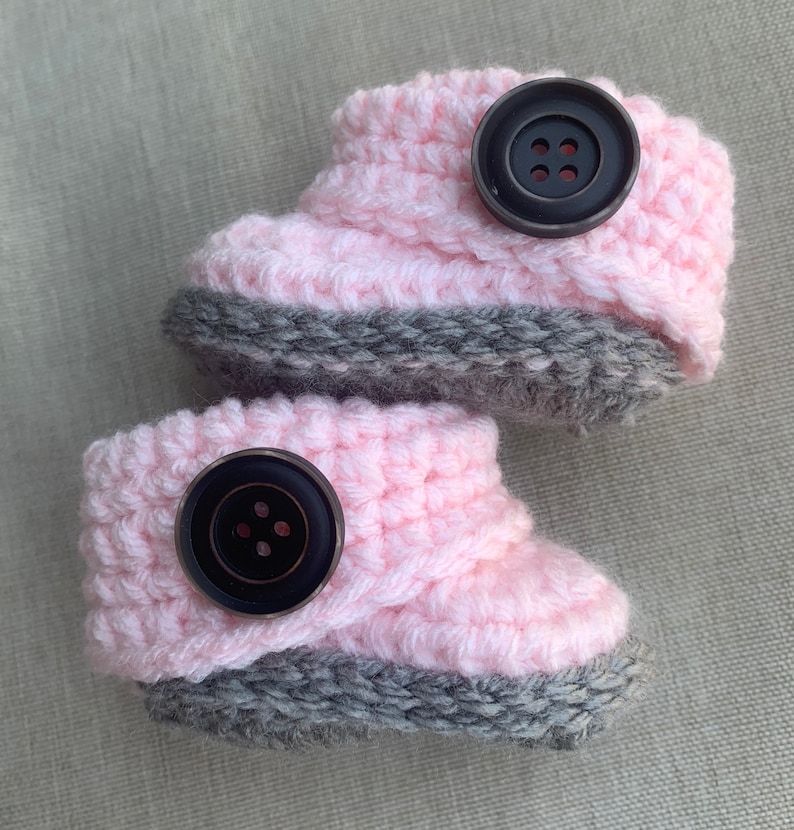 Op de afbeelding: Een paar roze en grijze gehaakte babyschoentjes met zwarte knopen.