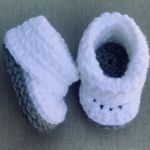 Op de afbeelding: Een paar witte gehaakte babyschoentjes met grijze bies. De schoentjes hebben een textuur en een klein zwart steekdetail aan de voorkant.