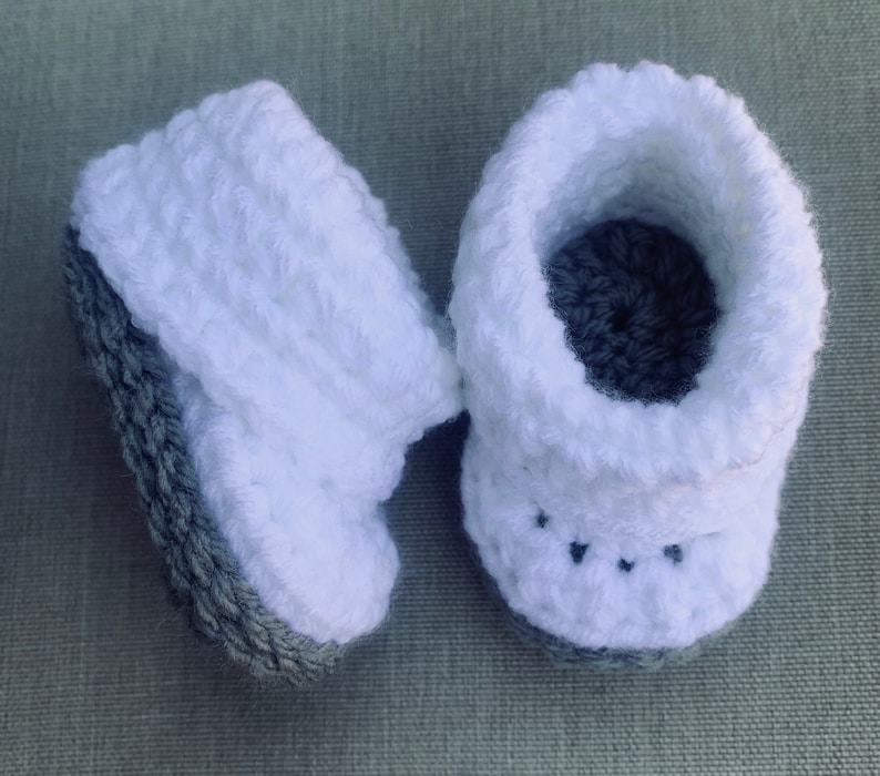 Op de afbeelding: Een paar witte gehaakte babyschoentjes met grijze bies. De schoentjes hebben een textuur en een klein zwart steekdetail aan de voorkant.