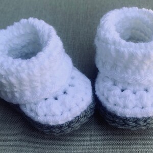 Op de afbeelding: Een paar witte gehaakte babyschoentjes met grijze bies.