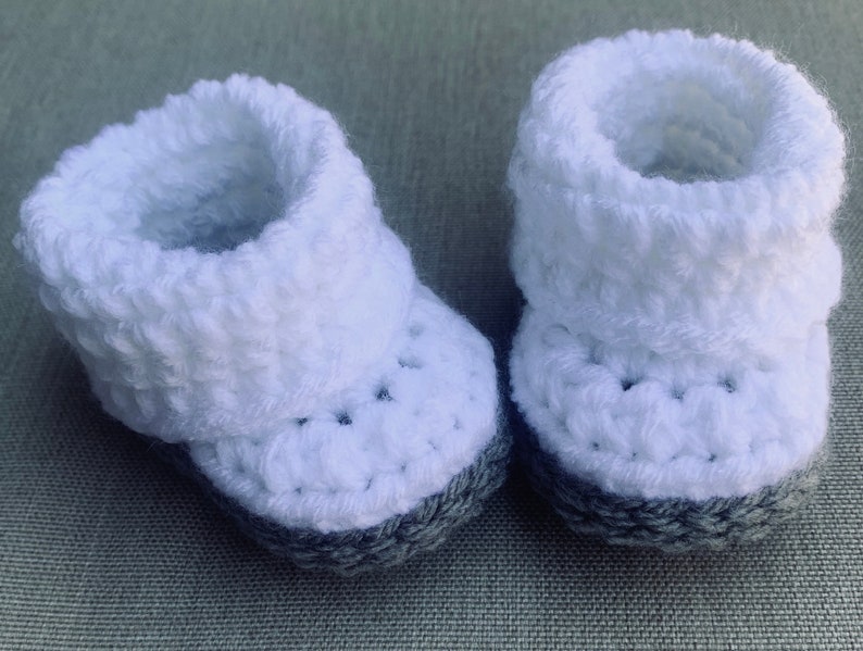 Op de afbeelding: Een paar witte gehaakte babyschoentjes met grijze bies.