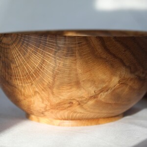 Op de afbeelding: Een houten kom met een natuurlijke houten afwerking. De kom heeft een brede, ronde vorm en een glad, gepolijst oppervlak. De houtnerf is zichtbaar en draagt bij aan de rustieke charme van de kom.