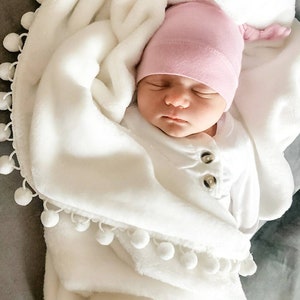 Op de afbeelding: Een pasgeboren baby draagt ​​een roze geknoopte muts en een wit rompertje en is gewikkeld in een wit deken met een witte pomponrand.