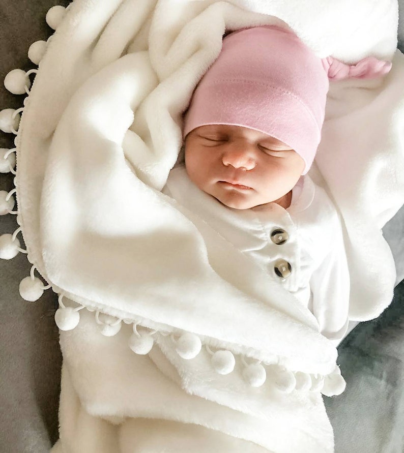 Op de afbeelding: Een pasgeboren baby draagt ​​een roze geknoopte muts en een wit rompertje en is gewikkeld in een wit deken met een witte pomponrand.