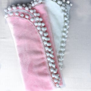 Op de afbeelding: Twee zachte dekens met witte pomponrand. Een deken is roze en de andere is wit.