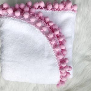 Op de afbeelding: Een witte badjas met een roze pompon rand. De badjas is dubbelgevouwen en de capuchon is zichtbaar.