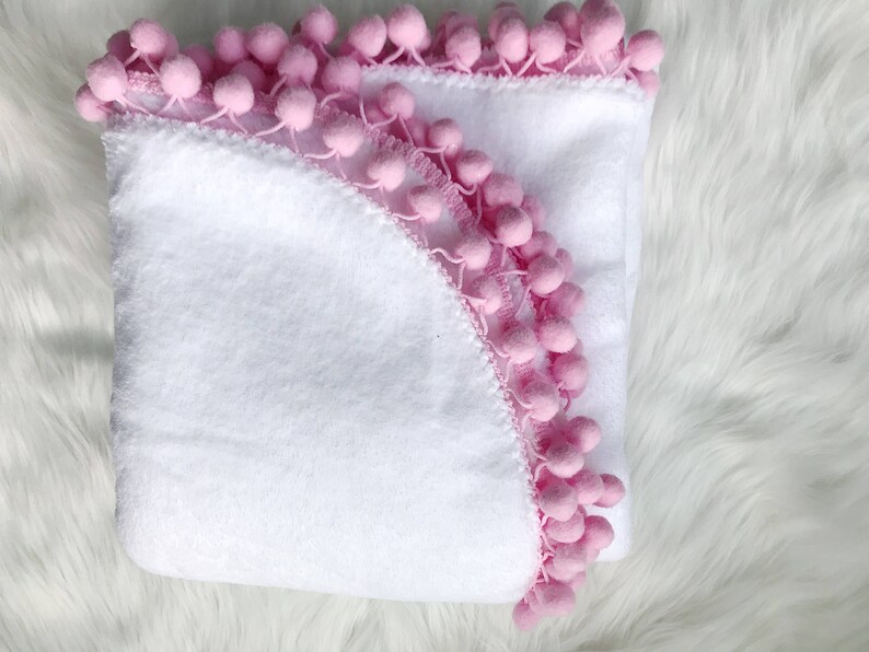 Op de afbeelding: Een witte badjas met een roze pompon rand. De badjas is dubbelgevouwen en de capuchon is zichtbaar.