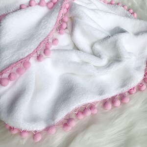 Op de afbeelding: Wit pluche deken met een roze pompon rand.