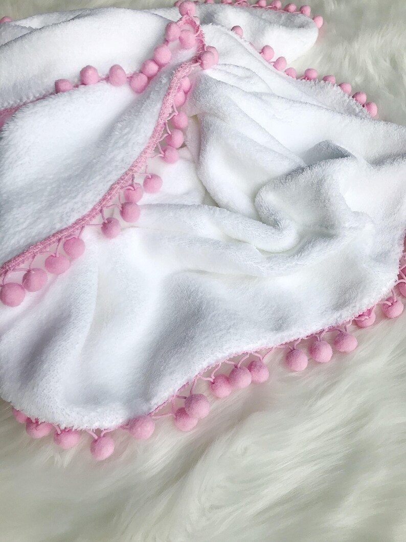 Op de afbeelding: Wit pluche deken met een roze pompon rand.