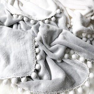 Op de afbeelding: Drie pluche dekens met witte pomponrand. De dekens zijn in de kleuren roze, wit en grijs.