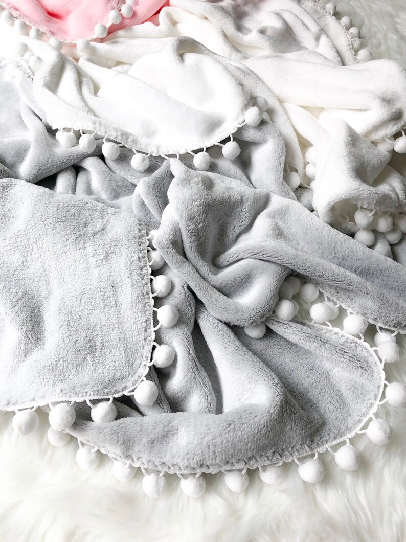 Op de afbeelding: Drie pluche dekens met witte pomponrand. De dekens zijn in de kleuren roze, wit en grijs.