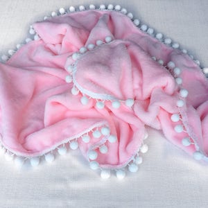 Op de afbeelding: Een zacht roze babydekentje met een witte pomponrand.