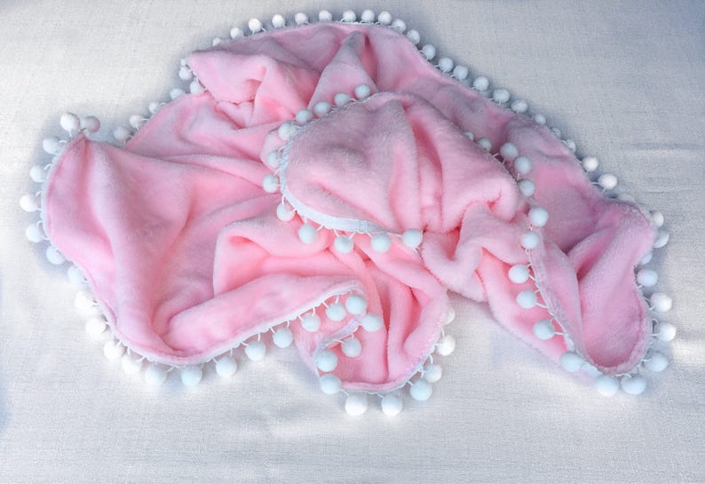 Op de afbeelding: Een zacht roze babydekentje met een witte pomponrand.