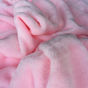 Op de afbeelding: Een zacht, roze pluche deken met een witte rand. Het deken is gevouwen en heeft een textuur.