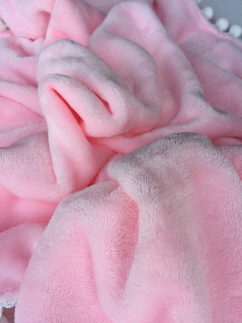 Op de afbeelding: Een zacht, roze pluche deken met een witte rand. Het deken is gevouwen en heeft een textuur.