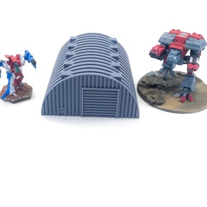 May include: A gray plastic model of a hangar with a corrugated metal door. The hangar is on a white surface. There are two miniature figures in the background, one red and one blue.