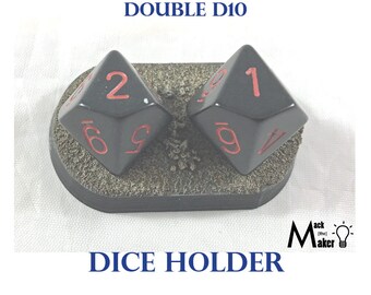 May include: A black plastic dice holder with two black ten-sided dice with red numbers. The dice holder has a textured surface and the dice are sitting in the holder. The text "DOUBLE D10 DICE HOLDER" is printed below the holder.