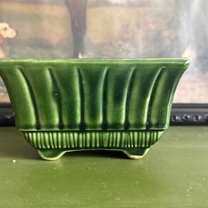 May include: A rectangular ceramic planter with a glossy dark green glaze. The exterior features vertical fluted ridges along the upper body and a textured, ribbed pattern near the base. The vessel is supported by four small, integrated feet.