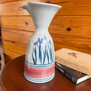 May include: A white ceramic pitcher with a flared rim and a narrow neck. The body features a hand-painted design of blue iris flowers with green stems above a horizontal band of soft pink and two thin blue stripes encircling the base.