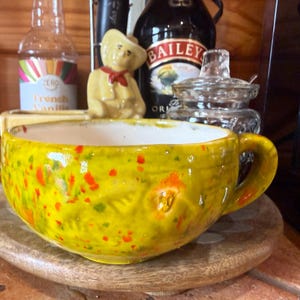 Puede incluir: Una taza de cerámica ancha y redonda con un esmalte amarillo brillante que presenta motas de pintura naranja y verde. La taza tiene un asa curva y lisa en el lado derecho y un interior blanco, mostrando una textura artesanal con hendiduras.