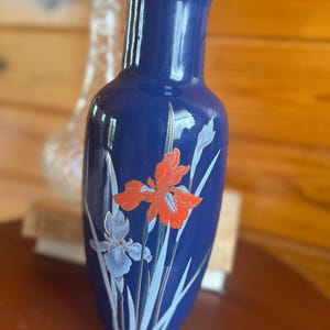 May include: A tall, cylindrical cobalt blue ceramic vase featuring a flared rim and a tapered base. The surface is decorated with a painted floral motif depicting vibrant orange and pale blue iris flowers with slender green leaves and stems.