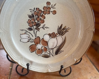 Puede incluir: Un plato redondo de cerámica con un esmalte blanquecino moteado y un borde marrón. El centro presenta un diseño floral pintado a mano en tonos marrones y blancos, que representa racimos de pequeñas flores y pétalos redondeados más grandes.