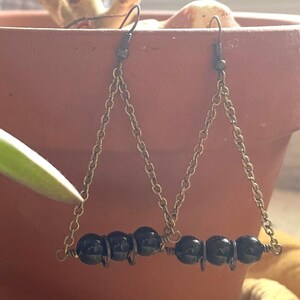 May include: A pair of black beaded earrings with a triangular design. The earrings hang from gold-colored chains and are displayed in front of a terracotta pot with a green plant.
