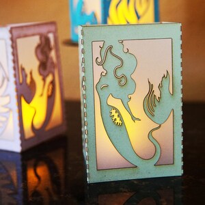 May include: A set of mermaid-themed lanterns, each with a unique design cut into the light green wooden panels. The lanterns are illuminated from within, creating a warm glow. Other lanterns feature seashell designs. The lanterns are approximately 15 cm tall.