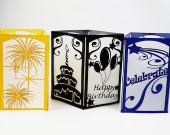 Könnte beinhalten: Drei dekorative Papierlaternen. Eine ist gelb mit einem goldenen Feuerwerksdesign, eine andere ist schwarz mit einer Geburtstagstorte und Luftballons, und die dritte ist blau mit dem Wort "Celebrate".