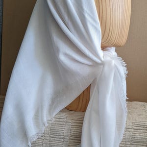 May include: A white scarf with fringed edges is draped over a wooden head form. The scarf is made of a lightweight fabric and is knotted at the base of the form. The background is a neutral beige.
