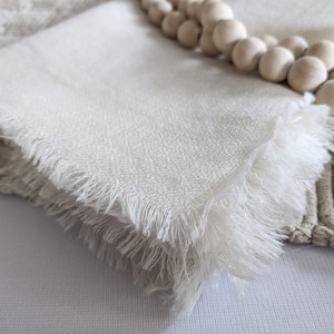May include: Off-white linen napkins with fringed edges, stacked on a textured surface. A strand of wooden beads and a woven placemat are also visible, suggesting a rustic or farmhouse aesthetic.