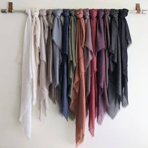 May include: A collection of 14 different colored scarves hanging on a rod. The scarves are all made of a soft, textured fabric and have a frayed edge. The colors include white, beige, gray, blue, green, brown, red, purple, and black.