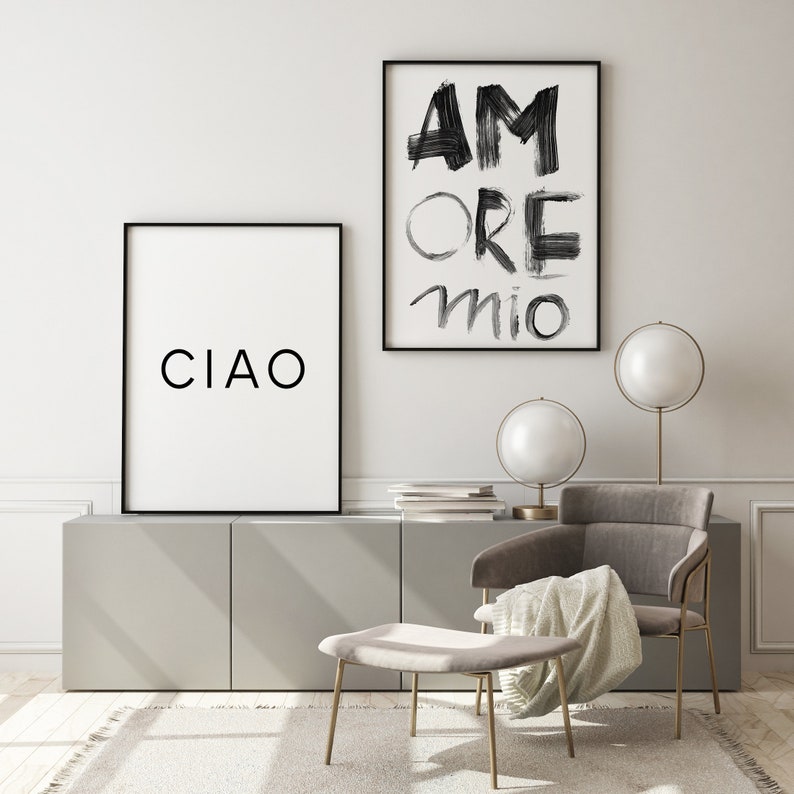 May include: Two framed black and white prints with the words "CIAO" and "AMORE MIO" hanging on a white wall above a gray cabinet. A gray armchair with a white throw blanket sits in front of the cabinet. A gold metal ottoman and two gold metal table lamps with white globe shades are also in the room.