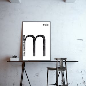 Könnte beinhalten: Ein schwarz-weißes Poster mit dem Buchstaben "m" in einer fetten, serifenlosen Schrift. Das Wort "mehr." ist in einer kleineren Schrift über dem Buchstaben gedruckt. Das Poster hängt an einer weißen Wand über einem schwarzen Tisch mit einem schwarzen Hocker.