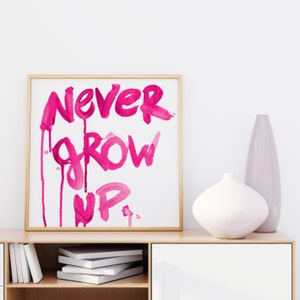 Puede incluir: Una impresi&oacute;n enmarcada con las palabras "NEVER GROW UP" escritas en pintura acuarela rosa. La impresi&oacute;n est&aacute; colgada en una pared blanca sobre un estante de madera con libros y jarrones.