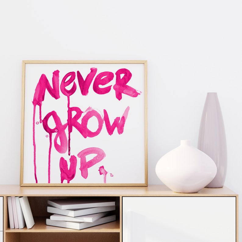 Puede incluir: Una impresi&oacute;n enmarcada con las palabras "NEVER GROW UP" escritas en pintura acuarela rosa. La impresi&oacute;n est&aacute; colgada en una pared blanca sobre un estante de madera con libros y jarrones.