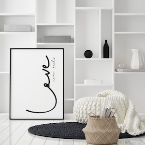 Könnte beinhalten: Ein gerahmter Schwarz-Weiß-Kunstdruck mit den Worten "love never fails" in minimalistischem Stil. Der Druck wird in einem hellen, modernen Raum mit weißen Regalen, einem schwarzen Teppich und einem Weidenkorb ausgestellt.