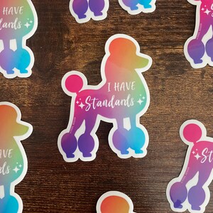 May include: A rainbow gradient sticker of a poodle with the text "I have standards" in white. The sticker is on a wooden surface.