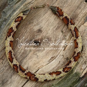 Pode incluir: Um colar de contas com um padrão manchado castanho e bege. O colar é circular e tem um fecho de bronze. O texto "Valentina Soldatova" está escrito em itálico na superfície de madeira.