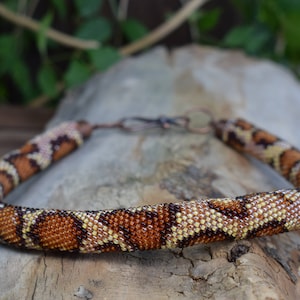 May include: A beaded necklace with a snake skin pattern in shades of brown, beige, and white. The necklace is made of small beads and has a clasp closure.
