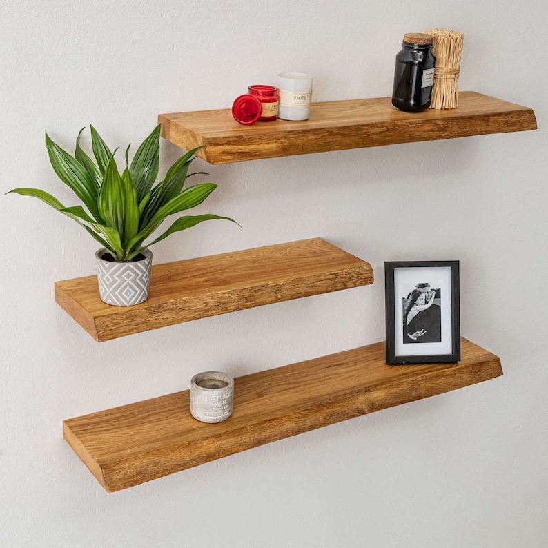 May include: Three wooden shelves with a natural finish, mounted on a white wall. The top shelf has two small candles and a jar with a cork lid. The middle shelf has a potted plant. The bottom shelf has a small candle and a framed photo.